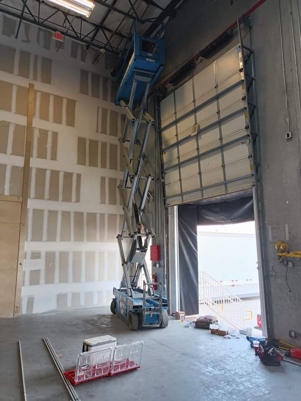 Scissor lift inside a warehouse near a large garage door, with tools and materials scattered.
