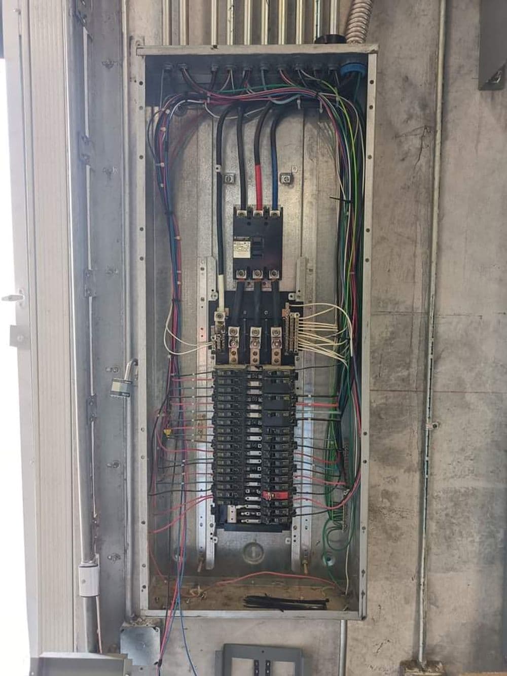 Electrical panel with multiple circuit breakers and exposed wiring on a gray wall.